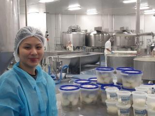 Woman wearing a hairnet next to tubs of cheese in a factory. There are factory workers behind her producing cheese.