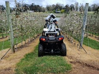 An all terrain vehicle with a high tech camera on the top driving in a flowering orchard.